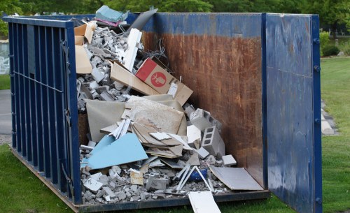 A large open-top industrial skip filled with a mixture of building debris and general waste, situated outdoors on a grassy area with a park or garden in the background. The contents include broken concrete slabs, plasterboard, wooden planks, and various construction materials, with some items spilling over the edges of the skip onto the surrounding ground. The skip has dark blue steel sides with rusted rebar markings visible on the interior surface and is positioned on a patch of grass near a paved walkway. The scene is well-lit with natural daylight, emphasizing the textures and colors of the waste materials. This image illustrates generic rubbish clearance, possibly in Norbiton or nearby South West London, with a focus on waste removal services such as those provided by Office Clearance Norbiton for commercial and domestic clearance projects.