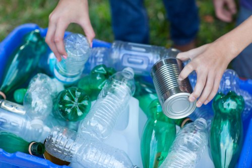 Close-up of a person's hands placing a plastic bottle and a tin can into a blue recycling bin filled with various clear and green plastic bottles and containers. The background shows a grassy outdoor area, suggesting a setting in Norbiton or nearby, with the focus on items related to waste collection and recycling. The person's hands are visible on the right side, with one hand holding the tin can by its sides and the other hand grasping a plastic bottle, indicating efforts to separate recyclable materials. The waste items exhibit different textures and finishes, with some bottles being smooth and glossy, while the tin can has a metallic sheen. The bin is partially visible, situated outdoors on a pavement or driveway, aligning with typical rubbish removal and recycling services offered by Office Clearance Norbiton.