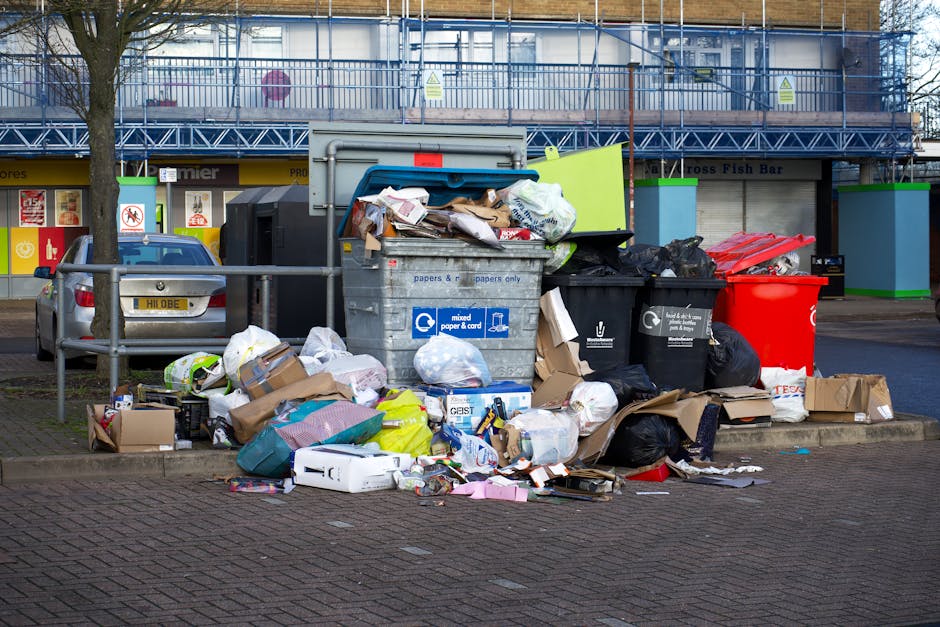 A collection of overflowing rubbish bins and waste materials situated on a paved parking area in front of a commercial building. The waste includes cardboard boxes, plastic bags, paper, and miscellane