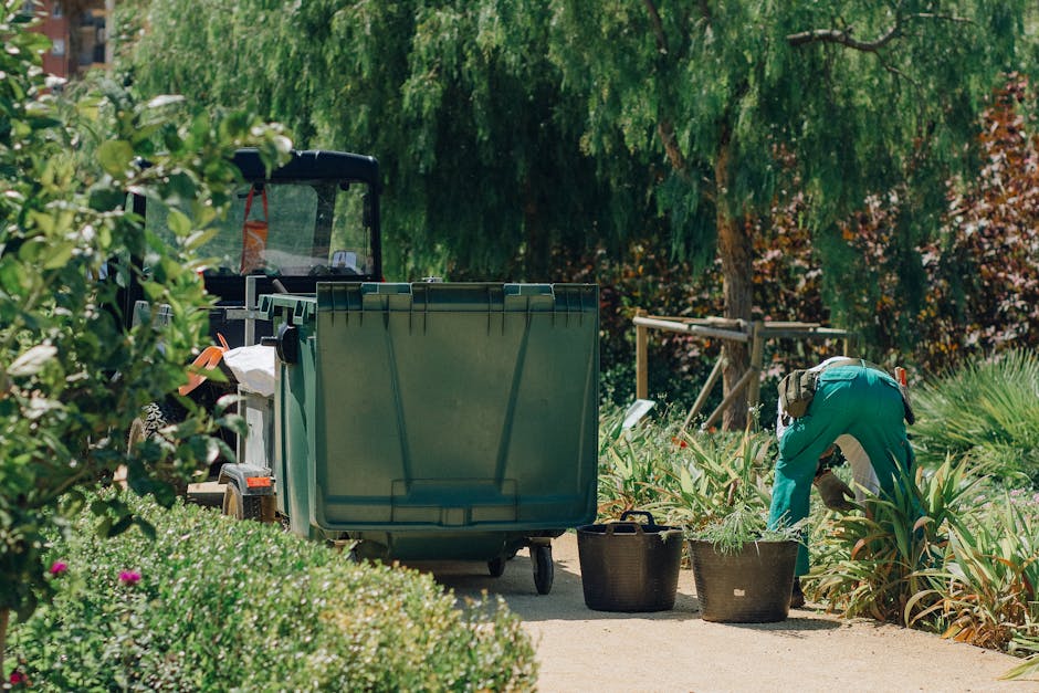 A residential street scene featuring four waste bins positioned on the pavement next to a low brick wall supporting a dense, green hedge. The bins are made of plastic with textured surfaces; three are