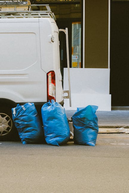 A large collection of waste and rubbish bags, cardboard boxes, and miscellaneous items piled on a paved urban sidewalk near a parking area. The rubbish includes black plastic bags, flattened cardboard