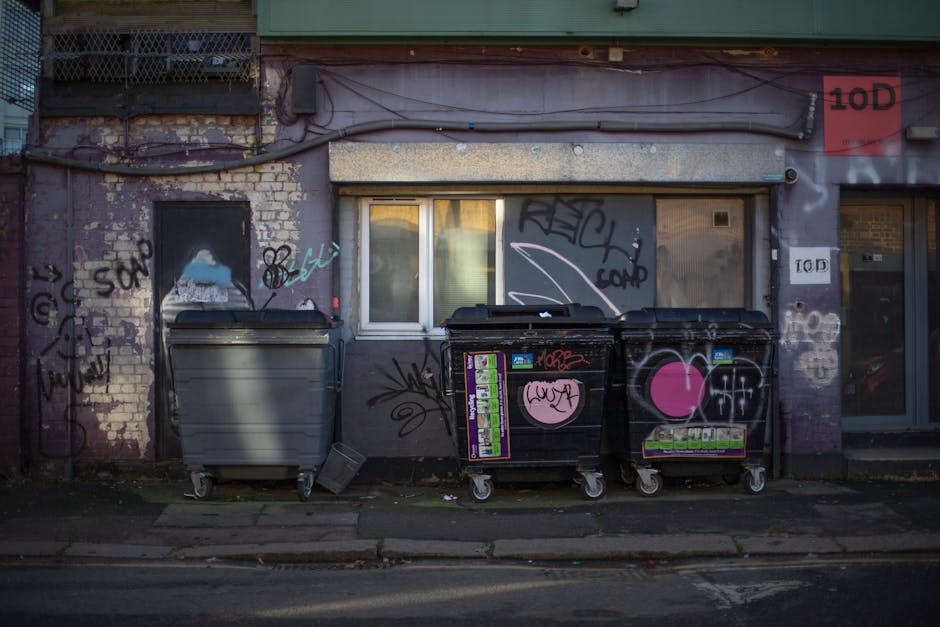 A large pile of mixed household rubbish and waste materials accumulated on a paved urban street corner, including a grey recycling bin overflowing with unflattened cardboard boxes, paper, and plastic